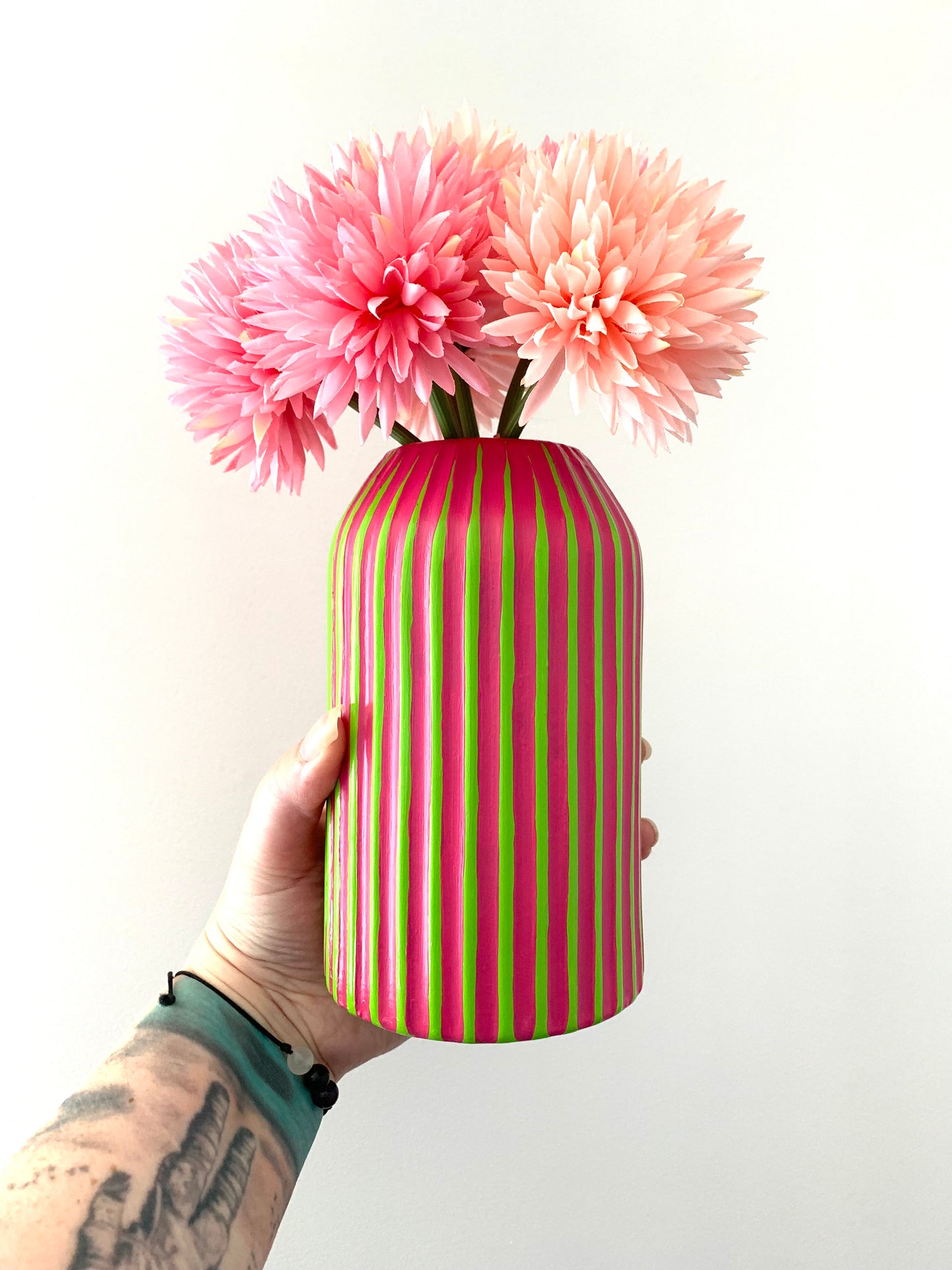Hand holding a green and fuchsia striped vase with pink flowers against a white background