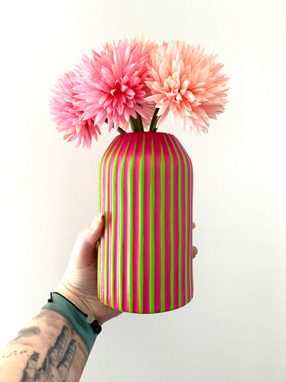 Hand holding a green and fuchsia striped vase with pink flowers against a white background