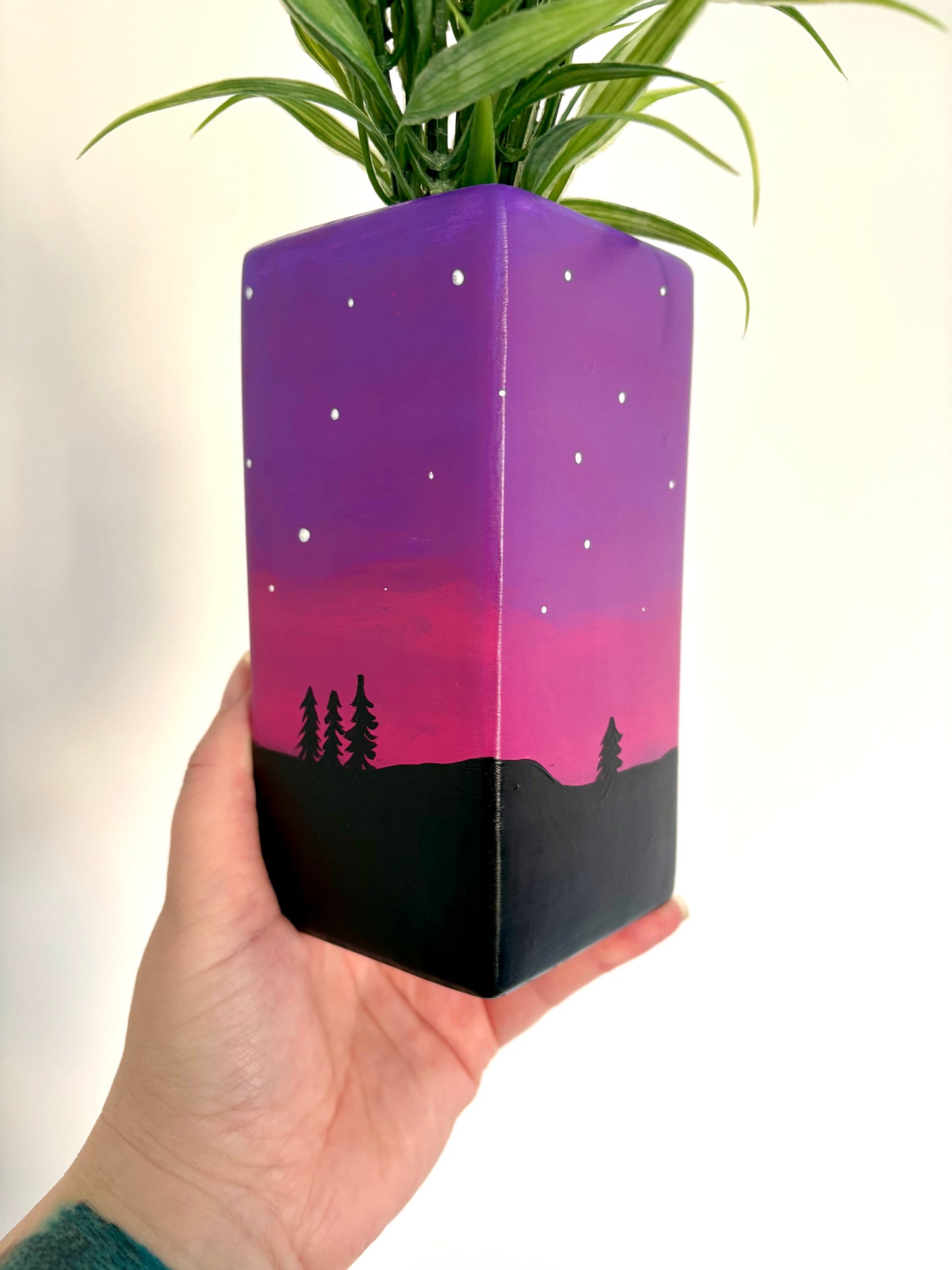 A square ceramic vase showing a hand painted night sky design featuring a purple and pink gradient, white stars, and a black tree silhouette