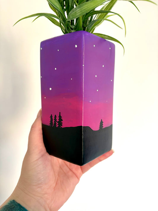 A square ceramic vase showing a hand painted night sky design featuring a purple and pink gradient, white stars, and a black tree silhouette