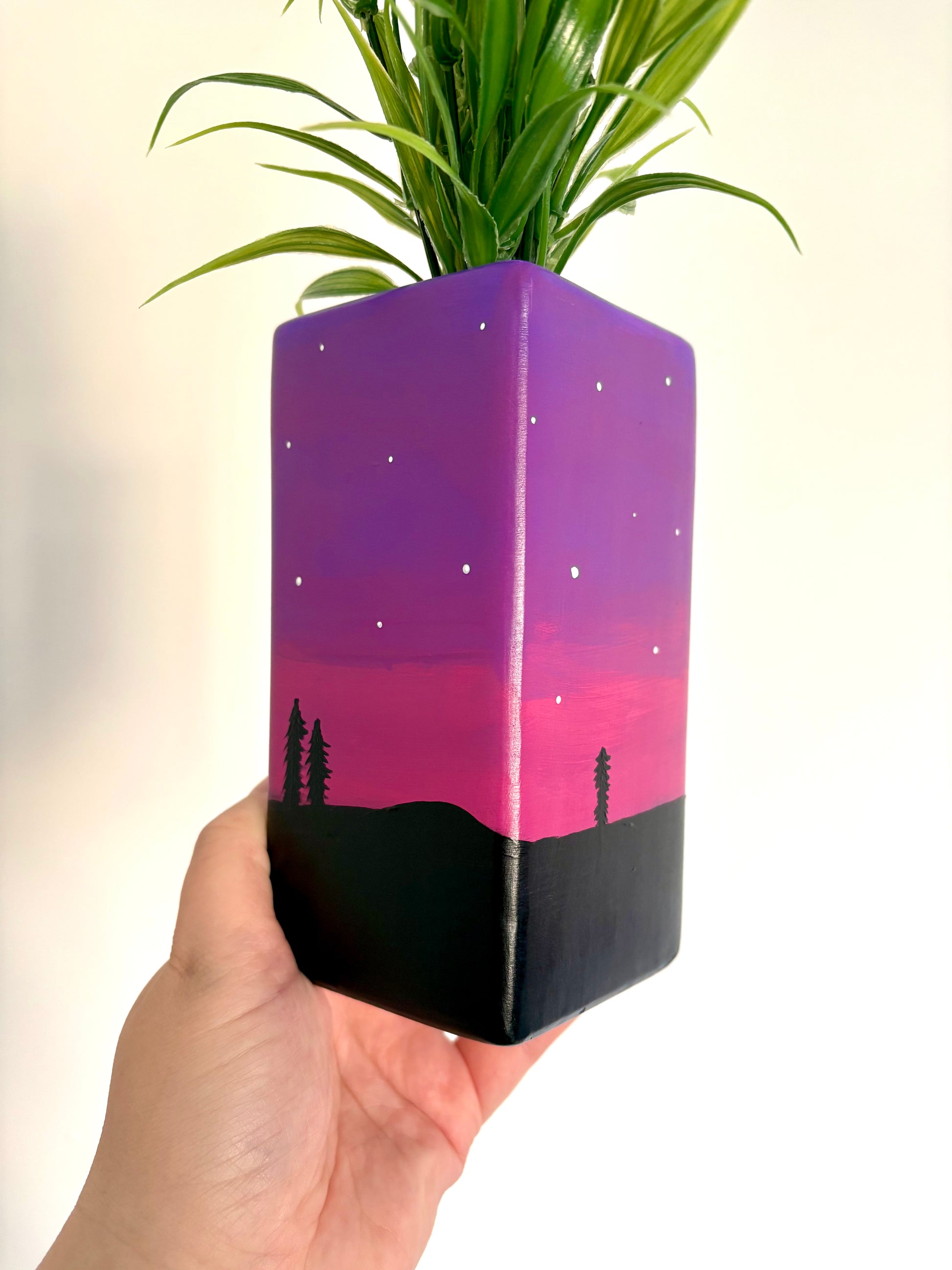A square ceramic vase showing a hand painted night sky design featuring a purple and pink gradient, white stars, and a black tree silhouette
