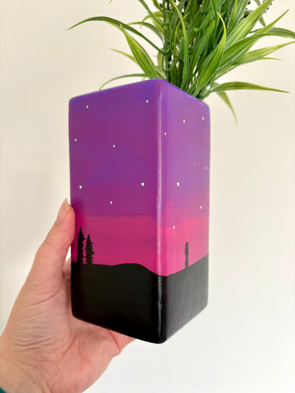 A square ceramic vase showing a hand painted night sky design featuring a purple and pink gradient, white stars, and a black tree silhouette